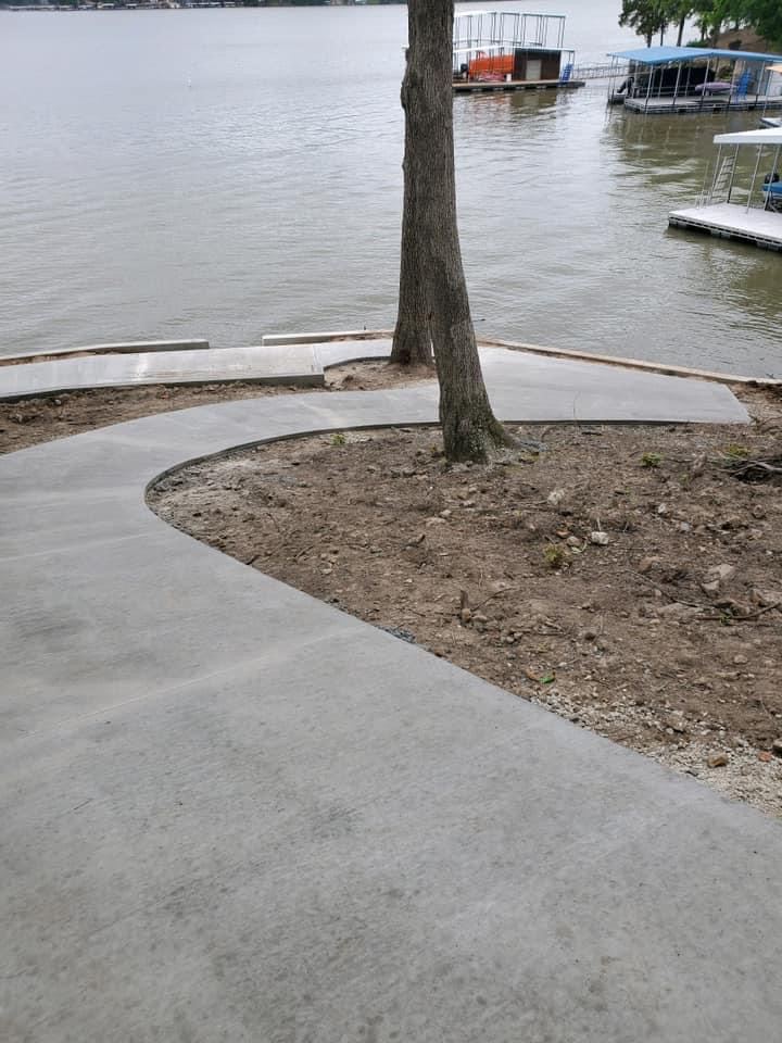 Curved lakeside sidewalk poured around a tree, boat docks behind